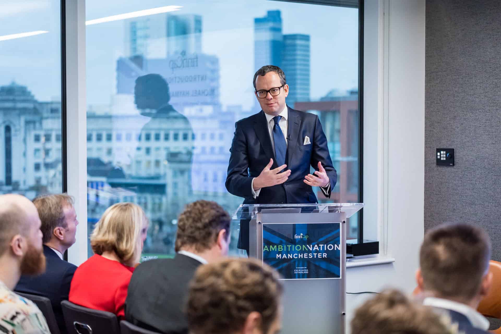 A man in a suit giving a speech in front of a group of people.