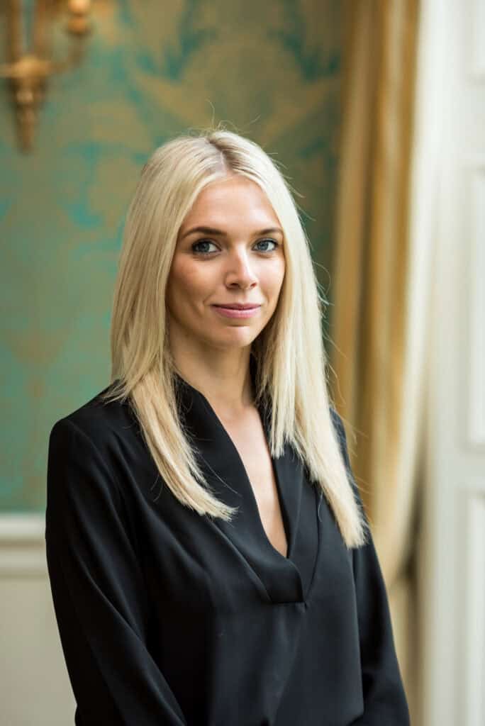 A blonde woman in a black shirt standing in front of an ornate room.