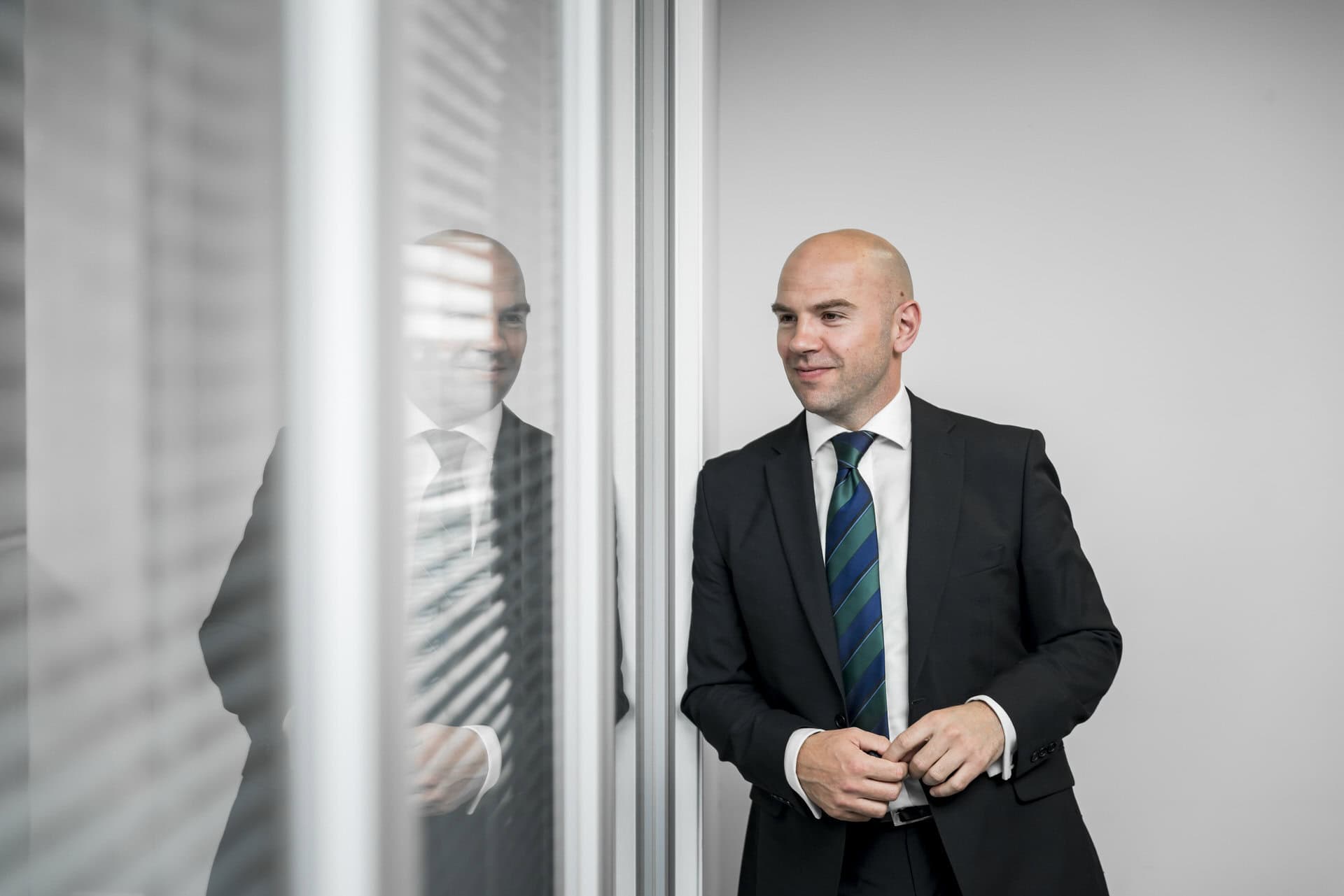 A bald man in a suit standing in front of a window.